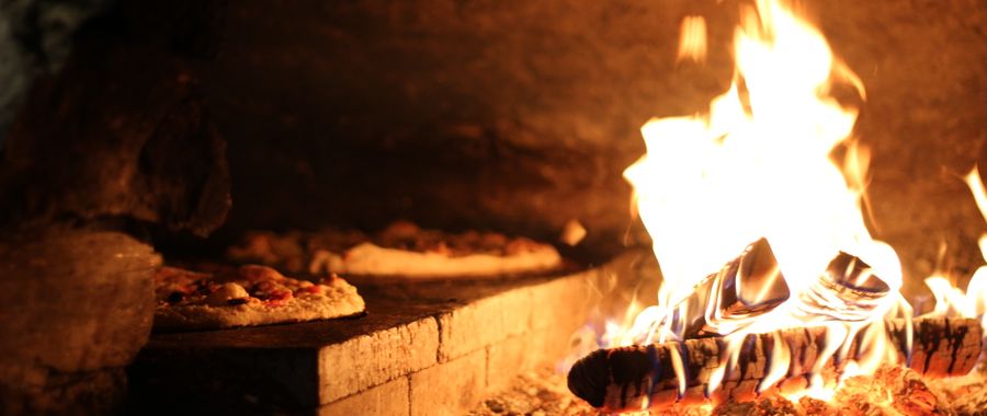 A rustic wood-fired refractory brick oven with pizzas near vibrant flames — the brick dome reflects radiant heat evenly across the cooking surface
